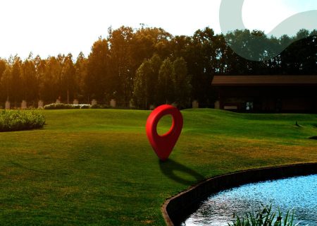 A large red map pin sculpture stands on a green lawn beside a pond. Trees and a building are in the background, with warm sunlight casting shadows.