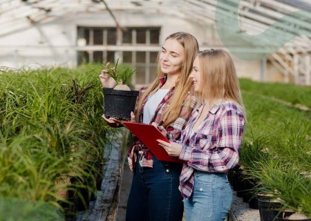 Two women in plaid shirts stand in a greenhouse, examining potted plants. One holds a plant, the other a red clipboard, both smiling and engaged.