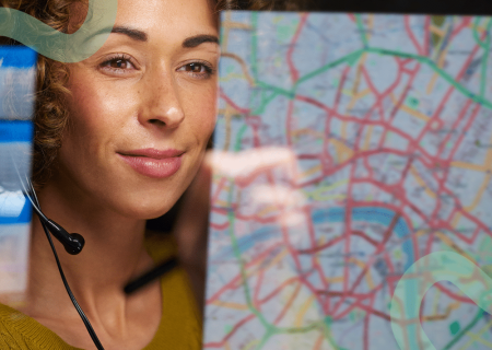 Woman with a headset smiles warmly, looking at a colorful city map reflected on a glass pane. The scene conveys focus and professionalism.