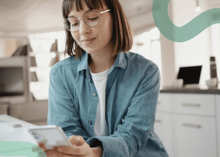 A woman with glasses, wearing a blue denim shirt, looks content while using a smartphone in a modern kitchen, conveying a relaxed and focused mood.