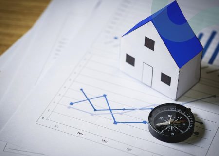A paper house with a blue roof sits on graphs with a blue line chart and a compass, symbolizing real estate trends and navigation.
