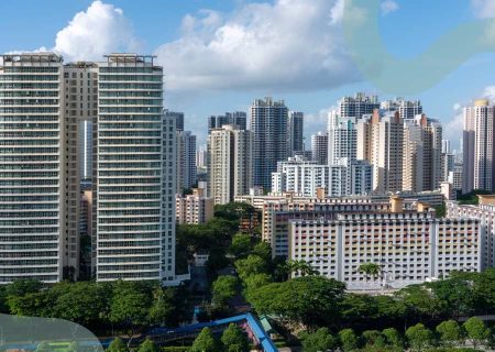 Skyline of a city with modern high-rise buildings under a blue sky with scattered clouds. Lush green trees are visible in the foreground.