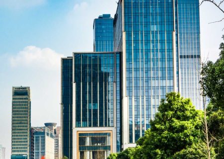 Modern skyscraper with a glass facade reflecting the blue sky, surrounded by lush trees and greenery, conveying a blend of urban and natural harmony.