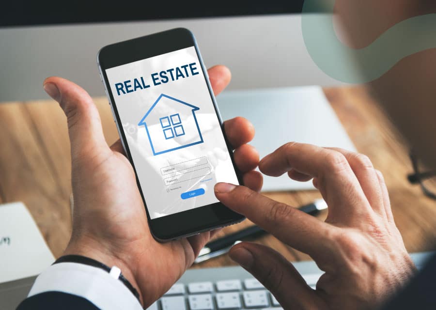 A person holding a smartphone showing a real estate app with a house icon and login screen. The setting is professional, conveying modern technology.