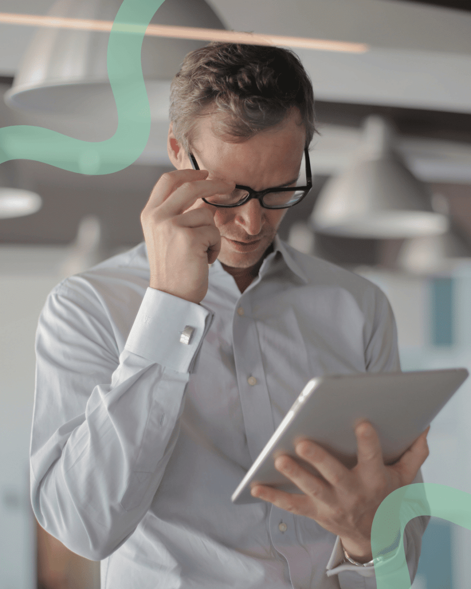 A man in a white shirt adjusts his glasses while focused on a tablet. He is in a bright office with pendant lights, conveying a professional tone.