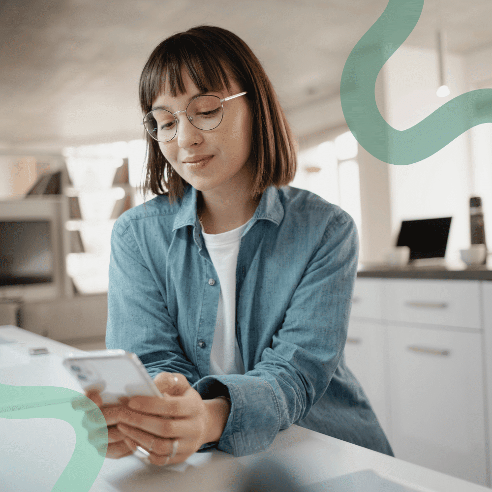 A woman with glasses, wearing a blue denim shirt, looks content while using a smartphone in a modern kitchen, conveying a relaxed and focused mood.