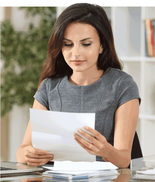 A woman in a gray dress sits at a desk, reading a document with a thoughtful expression. Behind her are plants and shelves with books, suggesting an office setting.