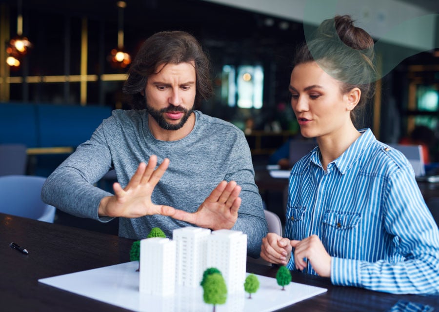 A man and woman discuss a white architectural model with small trees, appearing thoughtful and engaged. They are seated indoors, conveying a collaborative atmosphere.