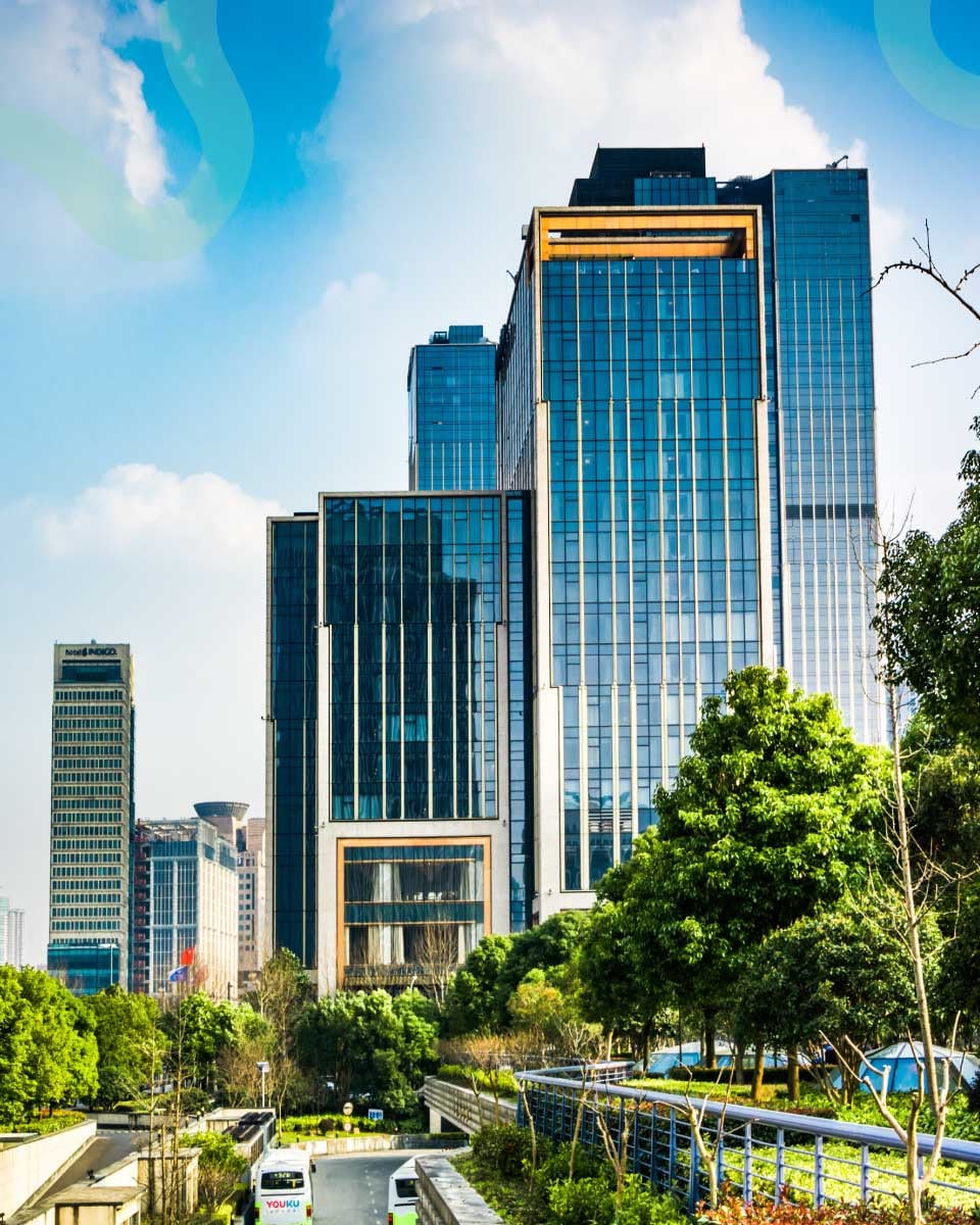 Modern skyscraper with a glass facade reflecting the blue sky, surrounded by lush trees and greenery, conveying a blend of urban and natural harmony.