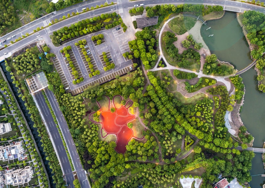 Aerial view of a vibrant park featuring a red, abstract-shaped pond surrounded by lush green trees. Nearby, winding paths and a parking lot are visible.
